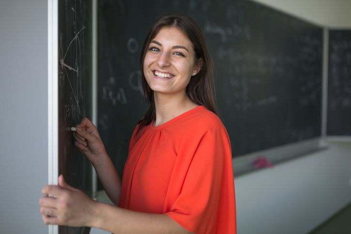 Ung, kvinnelig lærer skriver på en tavle mens hun ser i kameraet og smiler. Foto